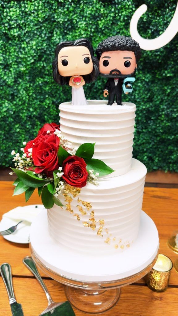 Simple, white, 2-tiered wedding cake with red roses and greenery floral placed throughout and wedding POP figures