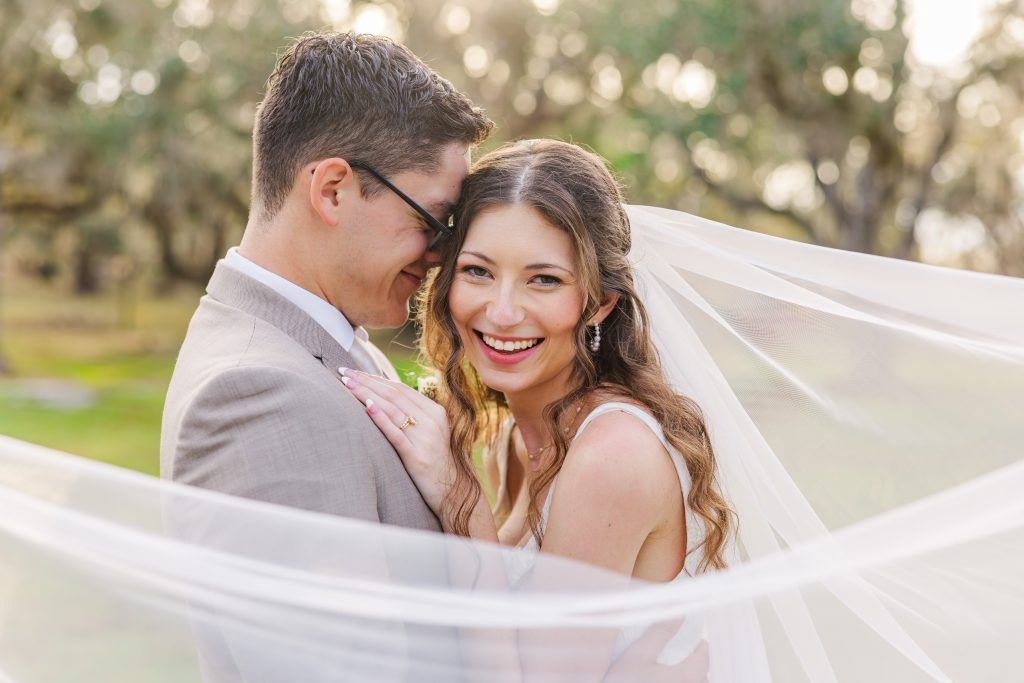 bride smiling at camera while groom smiles into her hair with the veil flowing
