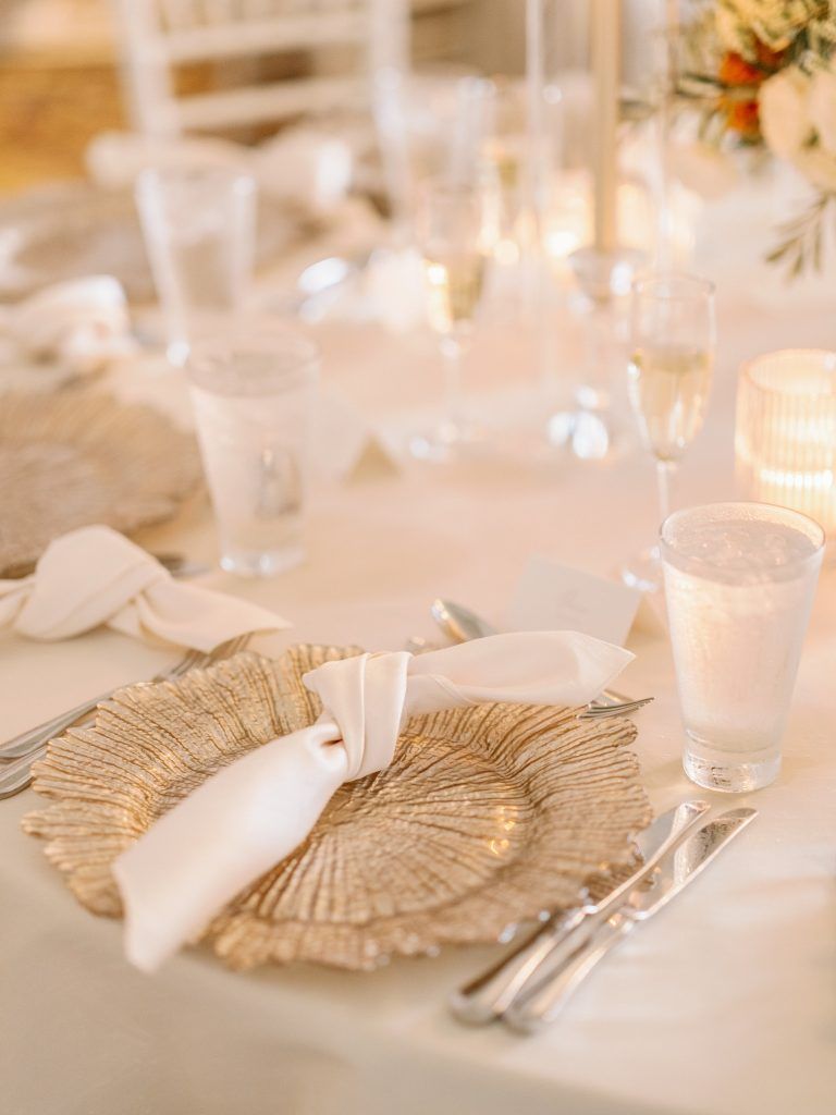 Elegant wedding place setting featuring a textured gold charger plate, a knotted ivory napkin, and polished silver flatware atop a soft ivory linen tablecloth. Surrounded by glowing candlelight and delicate glassware, this romantic tablescape is perfect inspiration for couples planning a luxurious, timeless wedding with neutral color palettes and refined details in Florida.