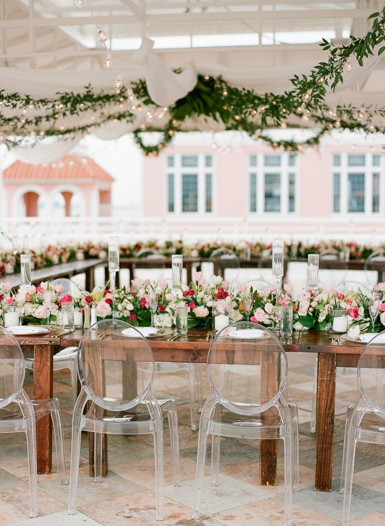 Romantic outdoor wedding reception featuring rustic wooden farmhouse tables decorated with lush pink and white floral arrangements, floating candles, and modern clear ghost chairs. Overhead, draped fabric, greenery garlands, and string lights create a whimsical, elegant ambiance—perfect inspiration for couples planning a garden wedding, rooftop reception, or blending rustic charm with modern wedding décor in Florida.