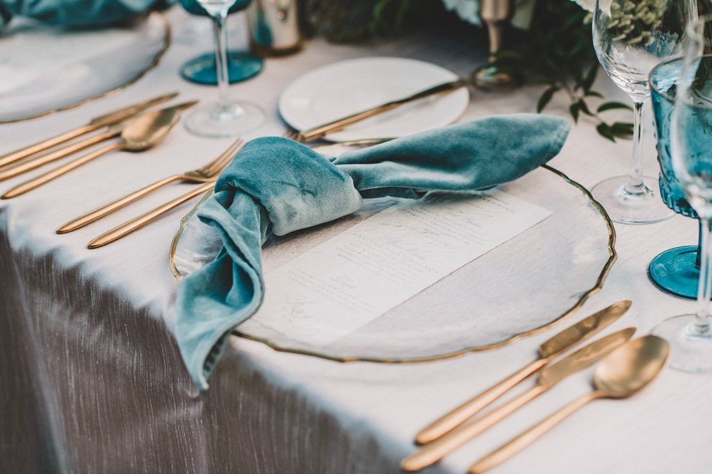 Luxurious wedding place setting featuring a clear glass charger with gold rim, modern gold flatware, and a soft velvet teal knotted napkin atop a textured linen tablecloth. The setting is accented by a printed menu card, blue glassware, and elegant greenery—perfect inspiration for couples planning a sophisticated wedding tablescape with jewel tones, velvet details, and chic modern décor in Florida.