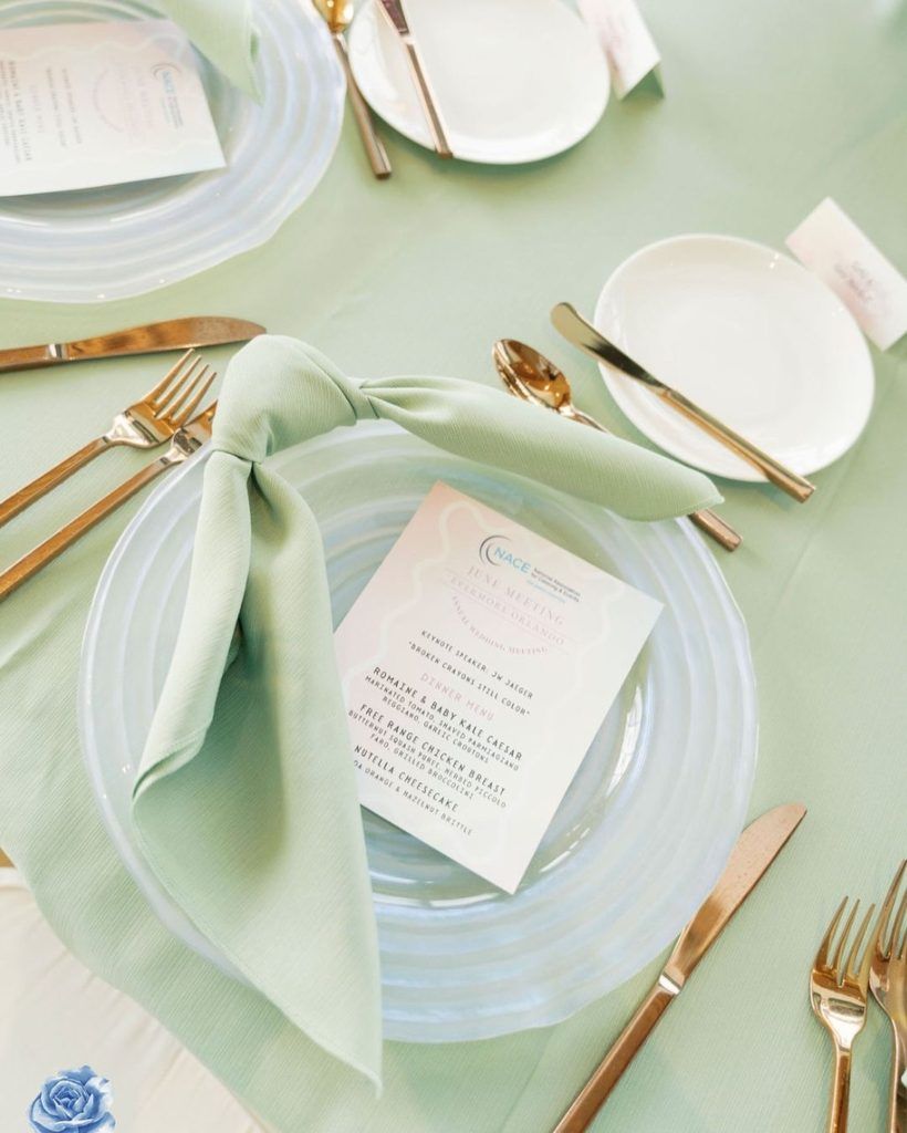 Modern wedding or event place setting featuring a clear glass charger plate, gold flatware, and a sage green knotted napkin atop a matching green linen tablecloth. The setting is completed with a printed menu card and simple white bread plate—perfect inspiration for couples seeking minimalist wedding table décor, trendy color palettes like sage green, and elegant gold accents for contemporary wedding receptions or styled events in Florida.