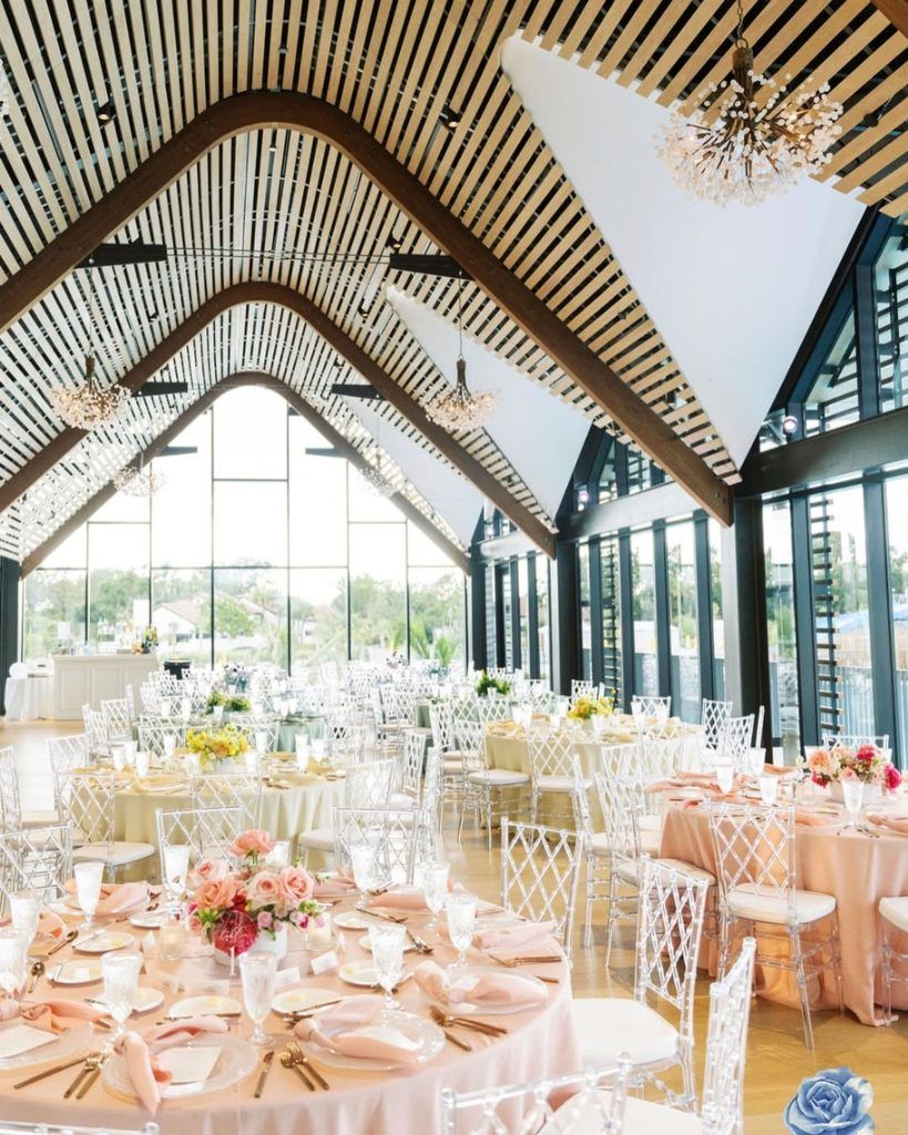 Modern wedding reception in a contemporary glass-walled venue featuring round tables with blush and champagne linens, clear acrylic chairs, and colorful floral centerpieces in shades of pink, peach, and yellow. The unique vaulted ceiling with wooden beams and statement chandeliers adds architectural elegance—perfect inspiration for couples seeking a bright, airy wedding venue with modern décor and romantic pastel accents in Florida.