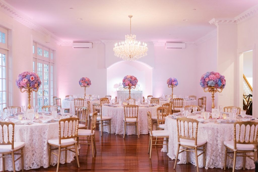 Chic wedding reception in a classic ballroom featuring round tables with textured ivory linens, gold chairs, and tall gold vases overflowing with colorful floral arrangements of pink, purple, and blue hydrangeas and roses. Soft pink uplighting and a sparkling chandelier create a romantic ambiance—perfect inspiration for couples planning a whimsical yet elegant wedding with bold florals and sophisticated décor in Florida.