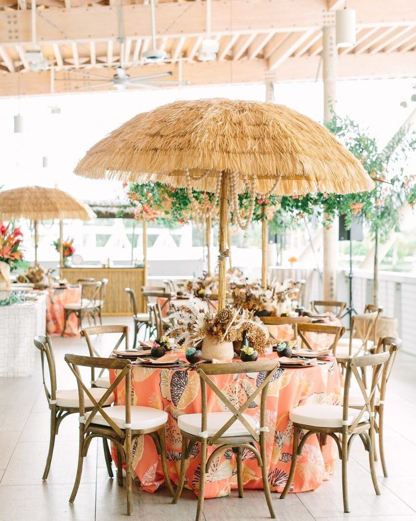 Tropical-themed wedding reception with round tables draped in bold coral and floral-patterned linens, accented by bamboo-style cross-back chairs and large thatched umbrellas with hanging beads. Centerpieces feature dried palm leaves and exotic florals, creating a fun, island-inspired atmosphere—perfect for couples planning a destination wedding, beach wedding, or luau-style celebration in Florida.