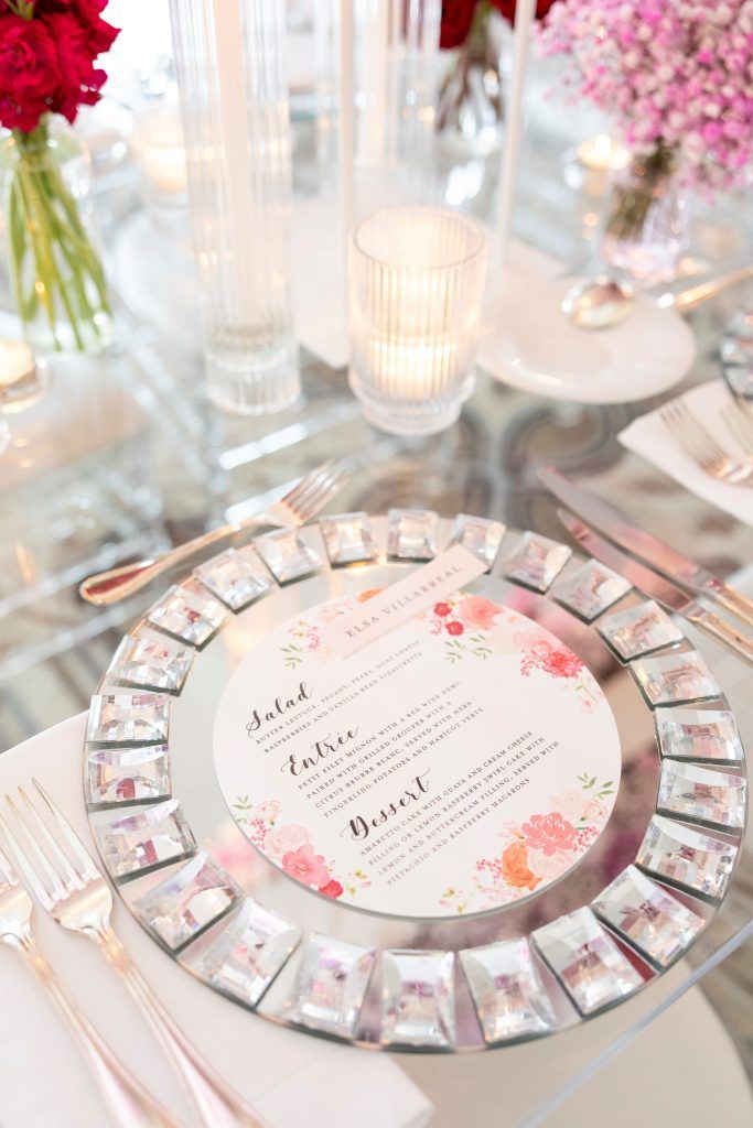 Elegant wedding table setting featuring a crystal and mirrored charger plate with a floral round menu card, surrounded by pink and red floral centerpieces and glowing candlelight on a glass table.