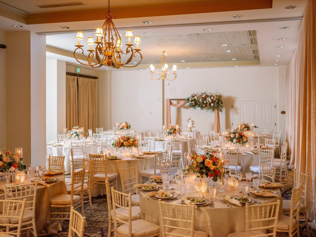 Elegant indoor wedding reception setup at a Florida wedding venue featuring round tables with ivory linens, gold chiavari chairs, romantic candlelight, and lush floral centerpieces in peach and cream tones. A decorated wedding arch and cake table are displayed in the background under classic chandeliers—perfect inspiration for couples planning a timeless and romantic wedding celebration.