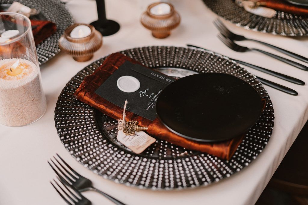Elegant wedding place setting featuring a textured black charger plate, rust-colored velvet napkin, black menu card with white wax seal, and black flatware.