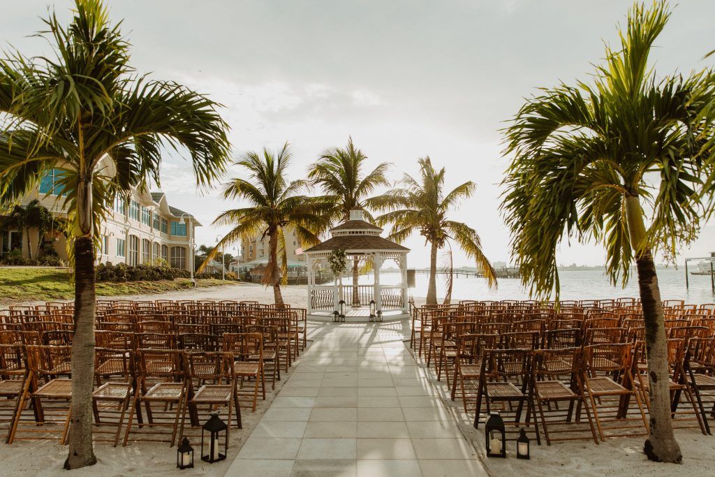 Romantic beach wedding ceremony with bamboo folding chairs, palm trees, and a white gazebo overlooking the ocean.