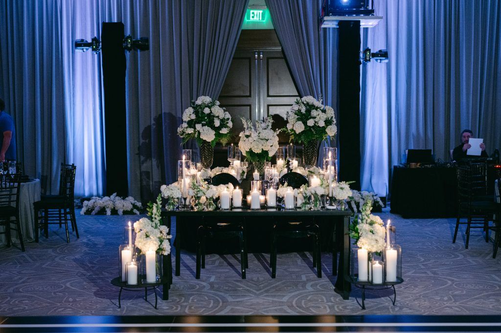 Luxurious wedding reception with black ghost chairs, dramatic white floral arrangements, and an abundance of candlelight on a black sweetheart table.