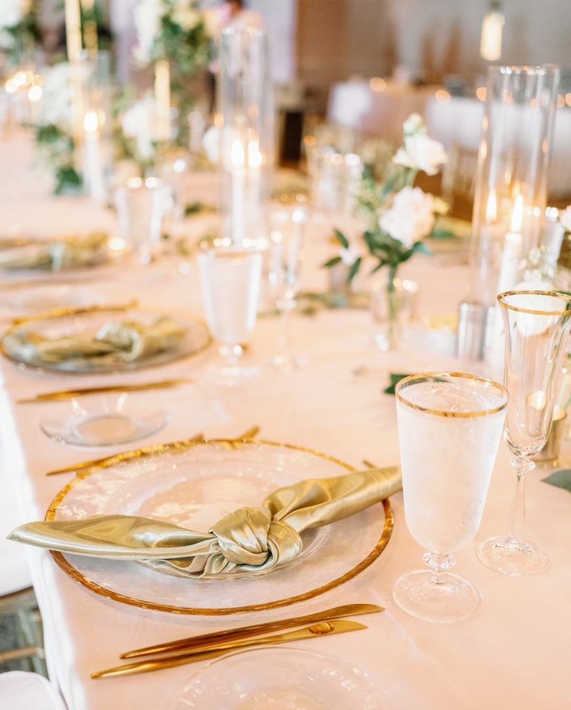 Elegant wedding reception place setting featuring a clear glass charger with a gold rim, a silky gold knotted napkin, gold flatware, and crystal glassware with gold trim. The table is accented with floating candles in tall glass cylinders and delicate white floral arrangements—perfect inspiration for couples planning a glamorous wedding with classic gold accents, romantic candlelight, and timeless décor in Florida.