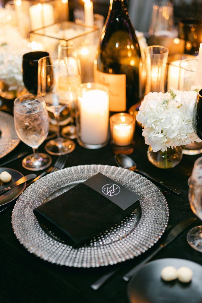 Elegant black and silver wedding reception table design featuring a black velvet napkin, silver beaded charger plate, white floral centerpiece, glowing candlelight, and black glassware for a dramatic and luxurious look.