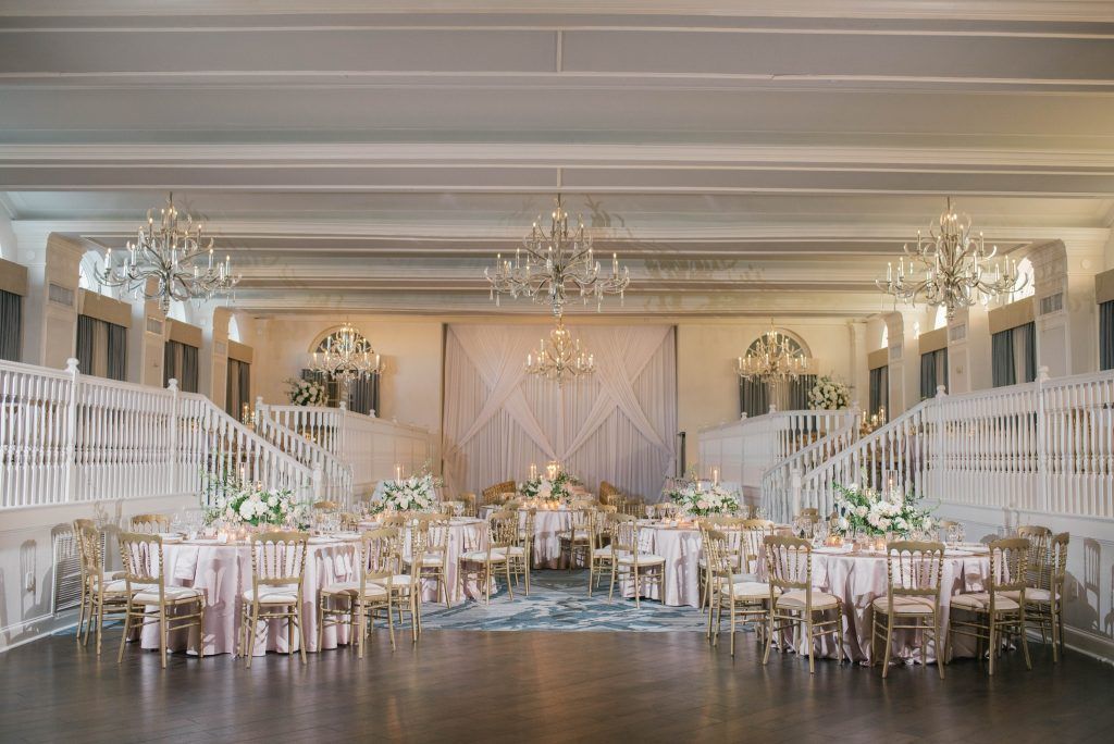 Elegant wedding reception at a luxury Florida wedding venue featuring gold chairs, blush pink satin table linens, lush white floral centerpieces, and grand chandeliers. The ballroom is designed with a split-level staircase and dramatic draping behind the head table, creating a timeless, romantic setting for engaged couples planning an upscale wedding.