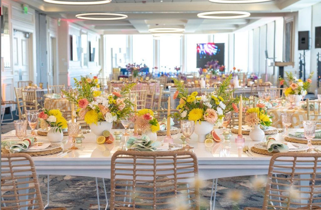Vibrant spring wedding reception featuring white tables with rattan chairs, colorful floral centerpieces of yellow, peach, and white blooms, and citrus fruit accents like lemons and grapefruits. Pastel napkins, pink glassware, and gold flatware add playful elegance to the cheerful tablescape—perfect inspiration for couples planning a bright, modern, and fresh spring or summer wedding in Florida.