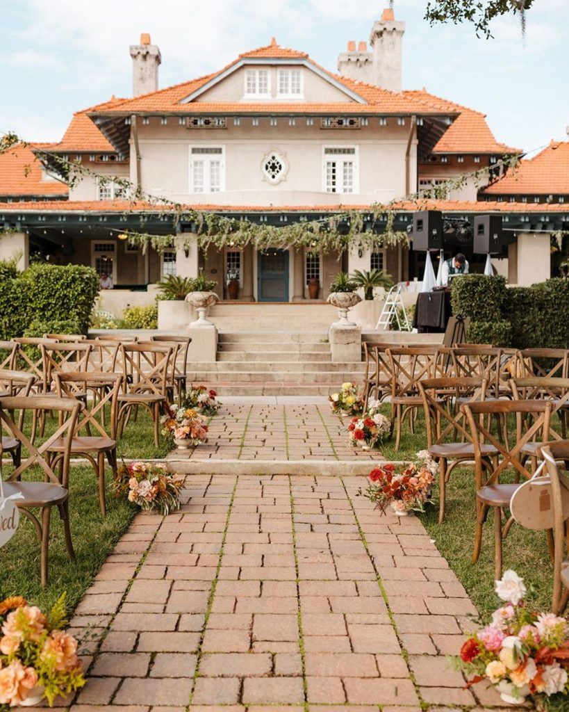 Rustic outdoor wedding ceremony set against a historic estate with terracotta roof tiles, featuring wooden cross-back chairs and a brick aisle lined with vibrant spring floral arrangements in shades of coral, red, and blush. Overhead greenery and string lights add to the romantic garden-inspired setting—perfect inspiration for couples planning a rustic elegant wedding, historic estate ceremony, or charming outdoor wedding in Florida.