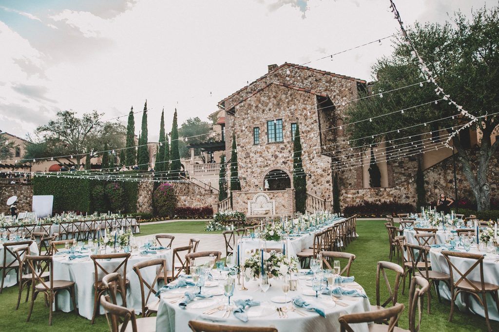Outdoor wedding reception at Bella Collina featuring Tuscan-inspired architecture, string lights, round and banquet tables with soft blue linens, natural wood cross-back chairs, and elegant white and blue floral centerpieces. A romantic, Italian-villa style setting perfect for a luxury wedding in Florida.