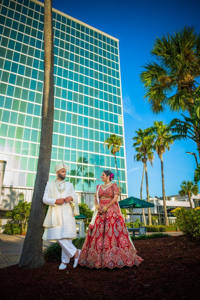 Bride and groom in traditional Indian wedding attire pose joyfully under the palm trees at DoubleTree by Hilton Orlando.