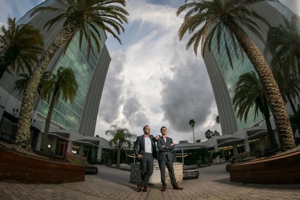 Two grooms in suits pose confidently beneath towering palm trees outside DoubleTree by Hilton Orlando.