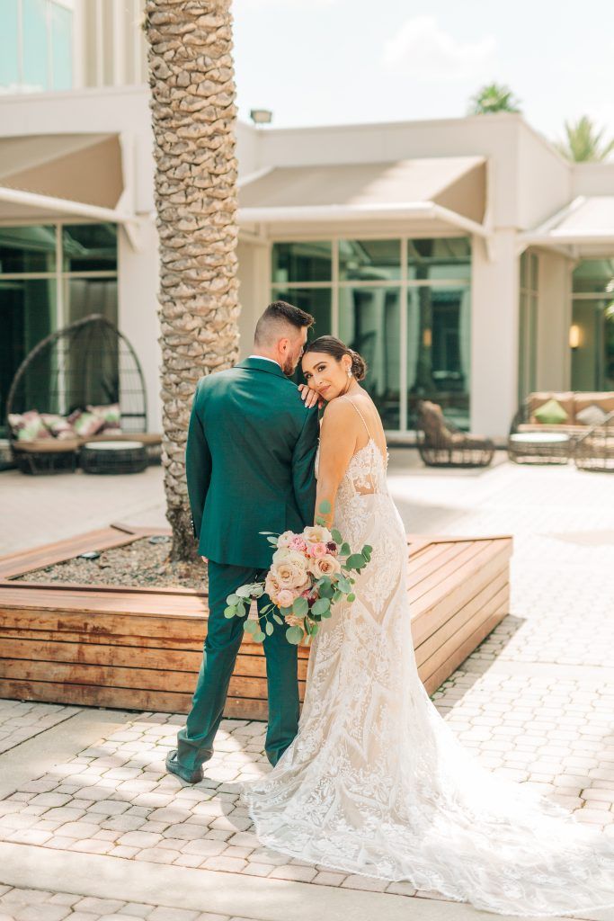 Bride in a lace wedding dress smiles over her shoulder while holding her groom’s hand outside DoubleTree by Hilton Orlando.
