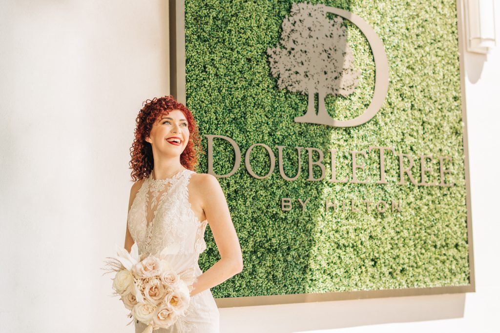 Smiling bride in a lace wedding dress poses in front of a green wall with the DoubleTree by Hilton Orlando sign.