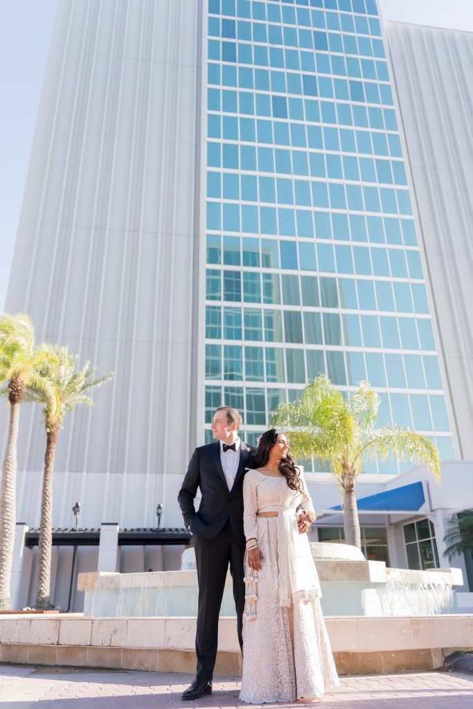 Bride and groom pose confidently outside DoubleTree by Hilton Orlando, with the hotel’s tall glass exterior behind them.