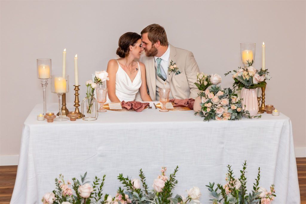 Bride and groom seated at their sweetheart table, surrounded by floral centerpieces and glowing candles, sharing a sweet kiss