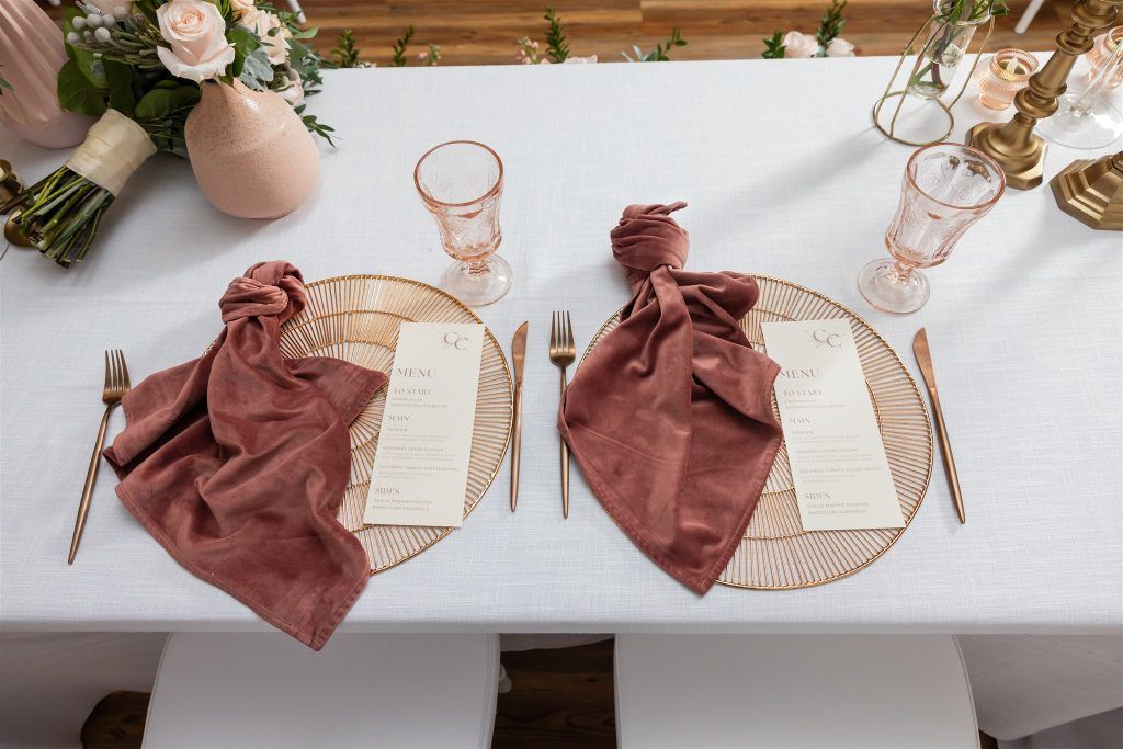Elegant sweetheart table styled with velvet mauve napkins, gold chargers, pink glassware, and printed wedding menus with floral arrangements