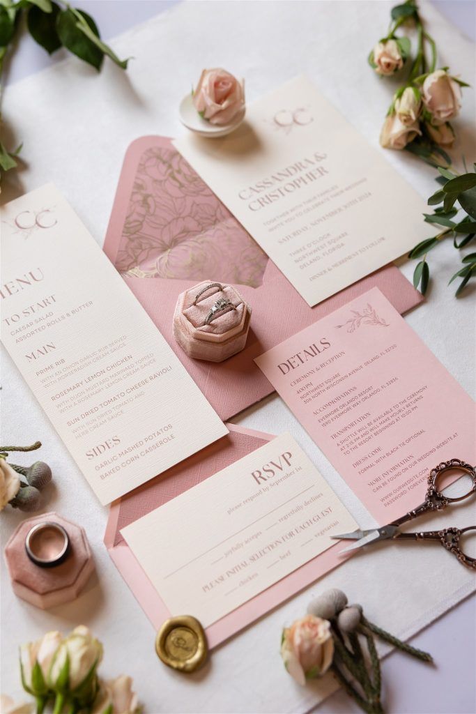 Flat lay of blush pink wedding stationery suite, featuring invitation, RSVP card, menu, and a velvet ring box with diamond ring