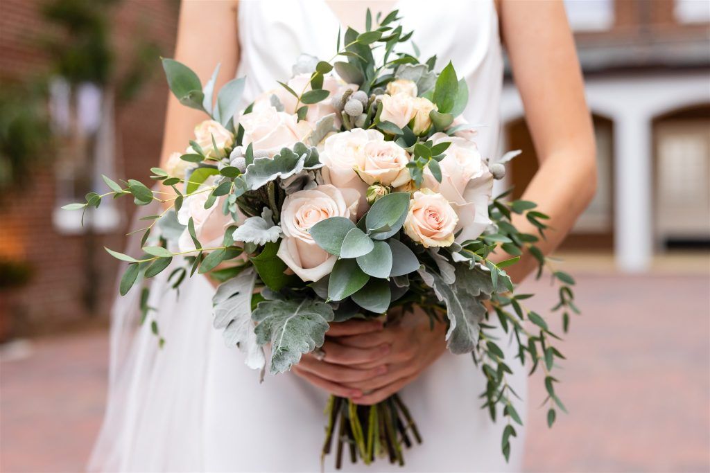 Bridal bouquet featuring blush and ivory roses, dusty miller, eucalyptus, and other greenery, held by the bride