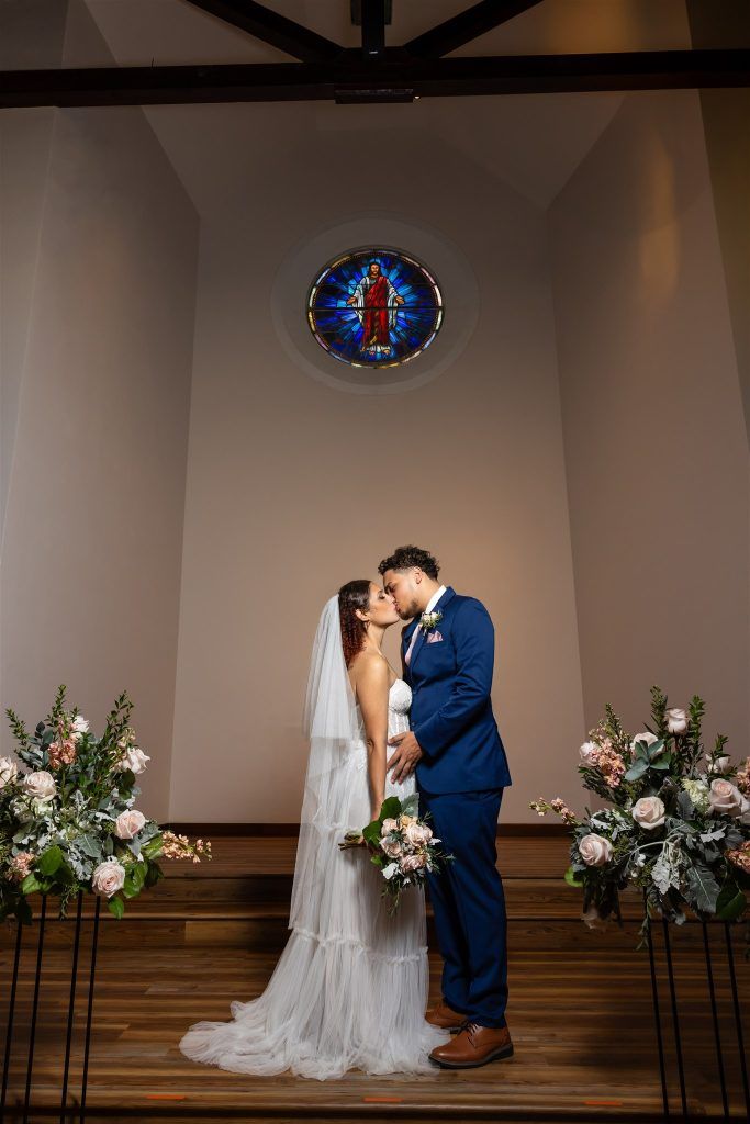 Bride and groom sharing a kiss under a stained glass window surrounded by tall floral arrangements in a romantic chapel.