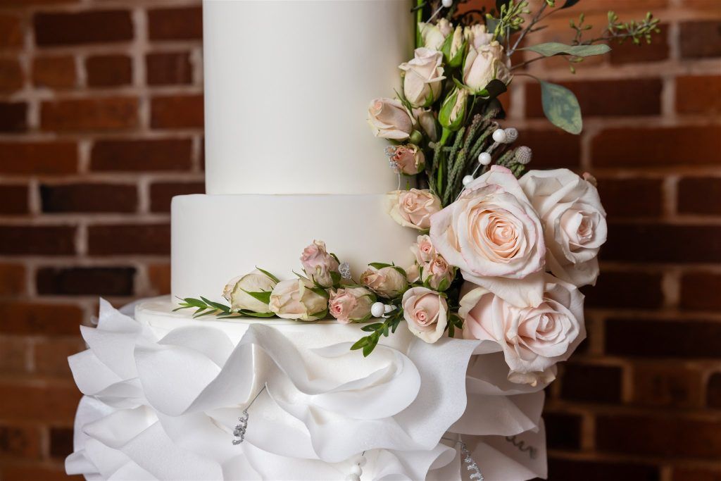Modern tiered wedding cake featuring white ruffled icing on the bottom tier and cascading blush roses and greenery