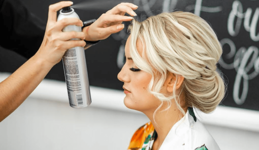 Kristy’s Artistry Design Team stylist applying hairspray to bride's elegant updo during wedding day hair and makeup prep.