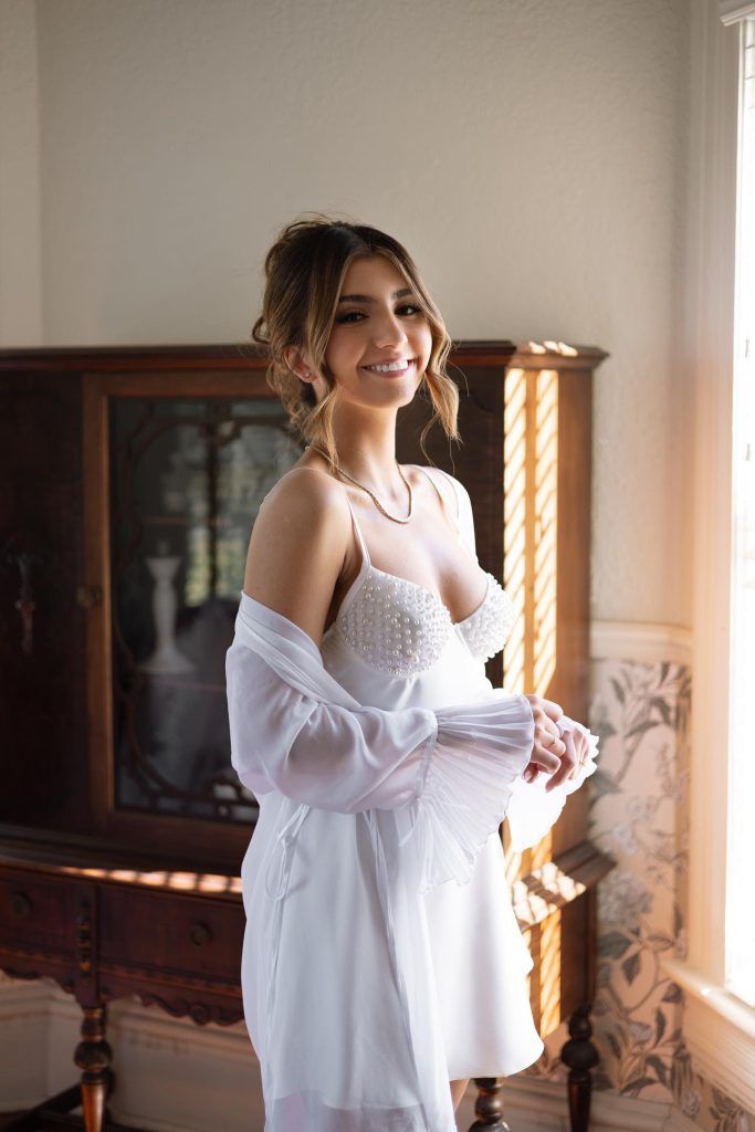 Bride wearing pearly-detailed white robe, styled with natural makeup and soft updo, standing by window with sunlight flowing in.
