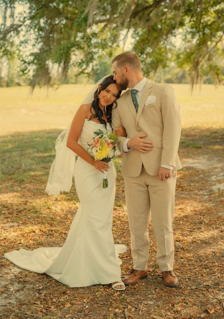 Bride in a strapless gown leans against her groom in a beige suit under large oak trees, holding pastel florals.