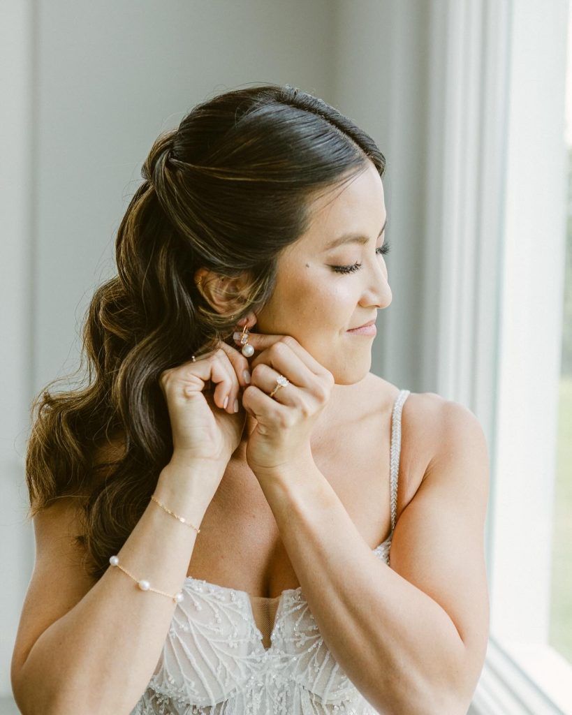 Bride with long brunette waves and soft glam makeup adjusting pearl earrings while standing near window before wedding ceremony.