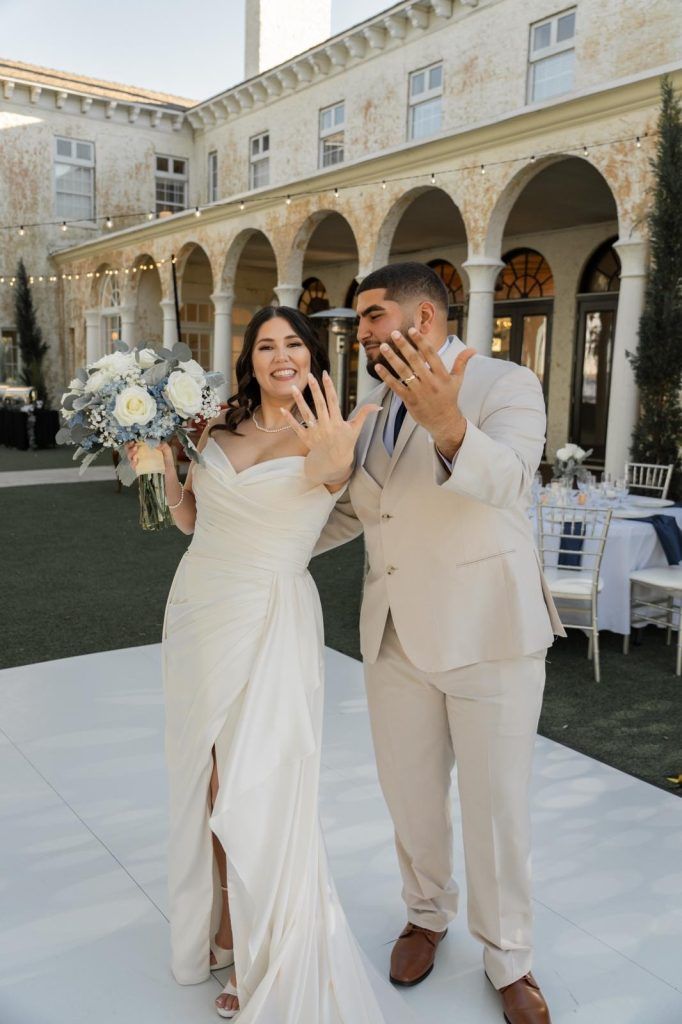 Newlywed couple smiling and showing rings at outdoor wedding, bride in off-shoulder white gown with natural glam hair and makeup.