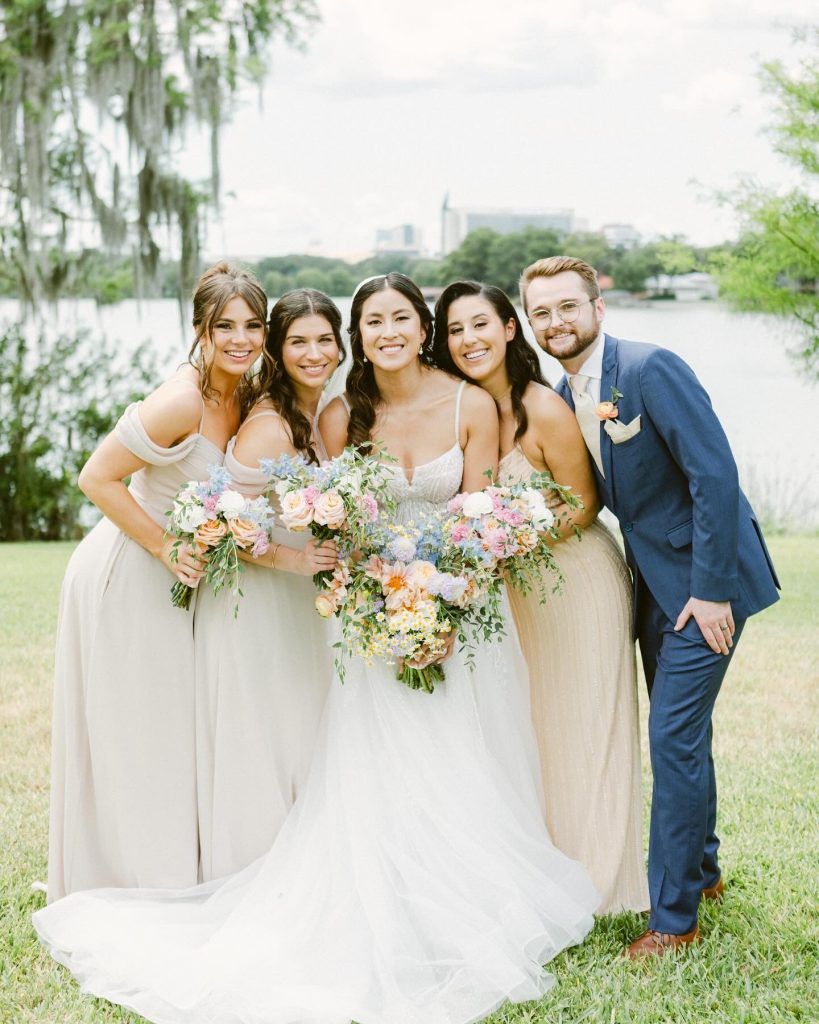 Bride with bridesmaids and groomsman outdoors, all smiling with colorful floral bouquets, showcasing The Glam Room hair and makeup looks.