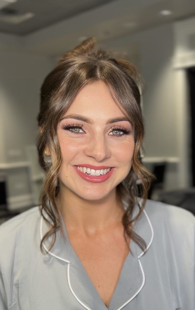 Close-up of a woman with glowing skin, pink lipstick, and loose curled updo smiling brightly in a gray pajama top.