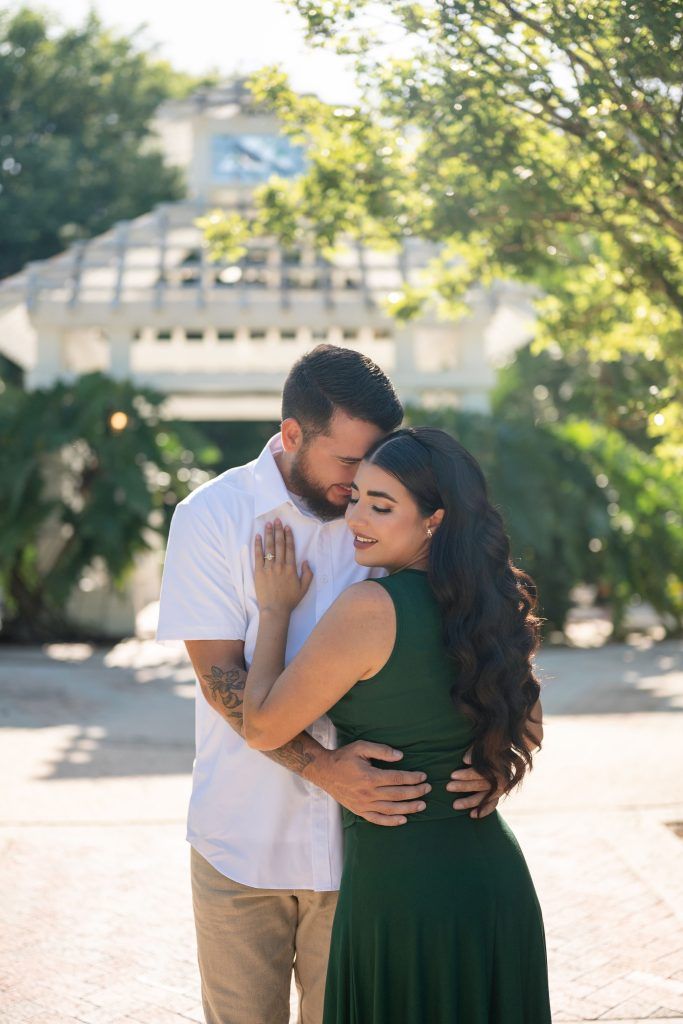 Couple embraces outdoors, the woman in a dark green dress with long waves resting against her partner in a white shirt.