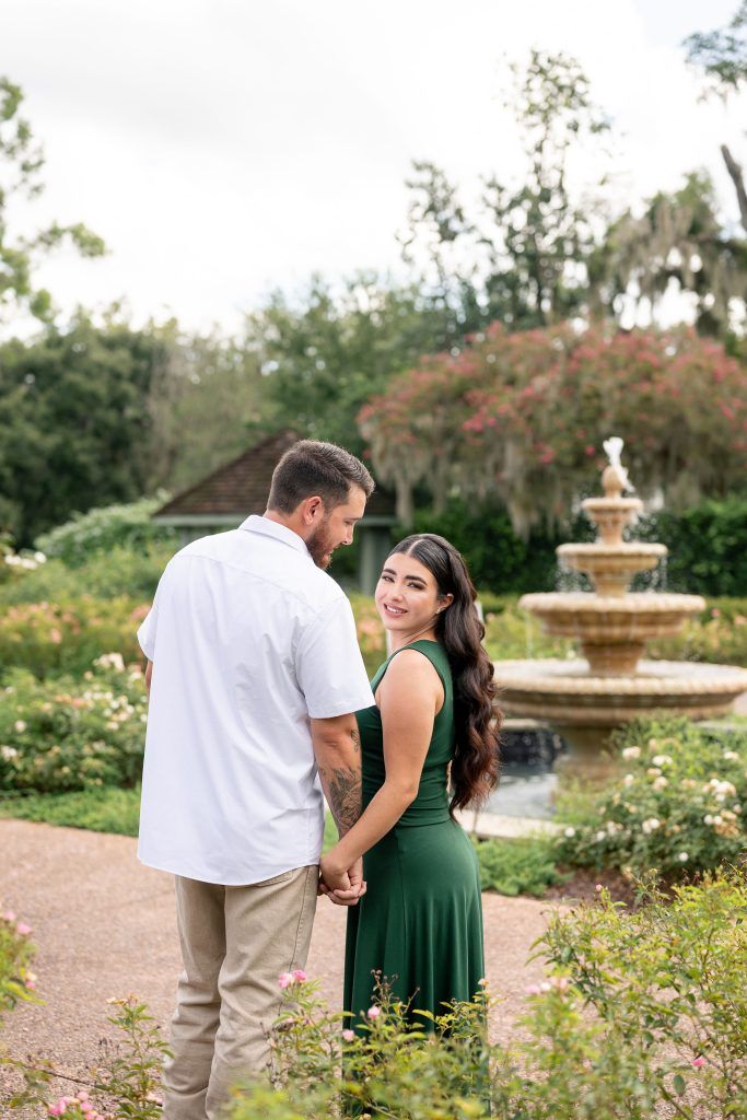 Couple holding hands in a garden with a fountain, the woman in a dark green dress smiling back over her shoulder.
