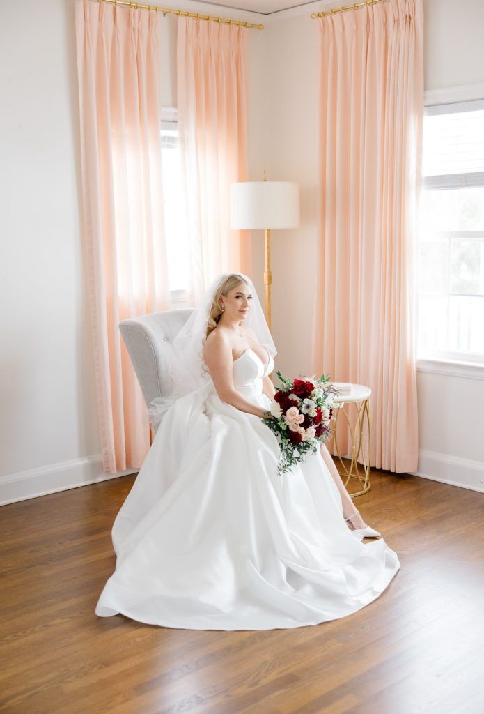 The bride sits elegantly in a chair holding a cascading bouquet of red, blush, and white flowers against a backdrop of peach curtains.