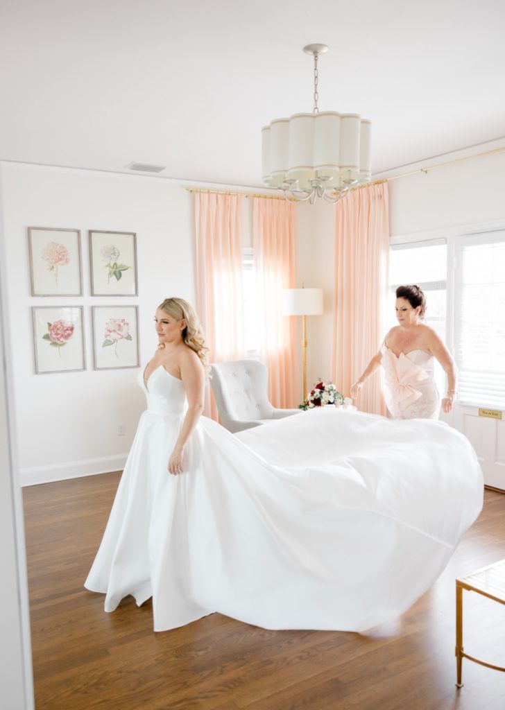 The bride walks across a room with her mother behind her, holding her voluminous white gown to keep it perfectly arranged.