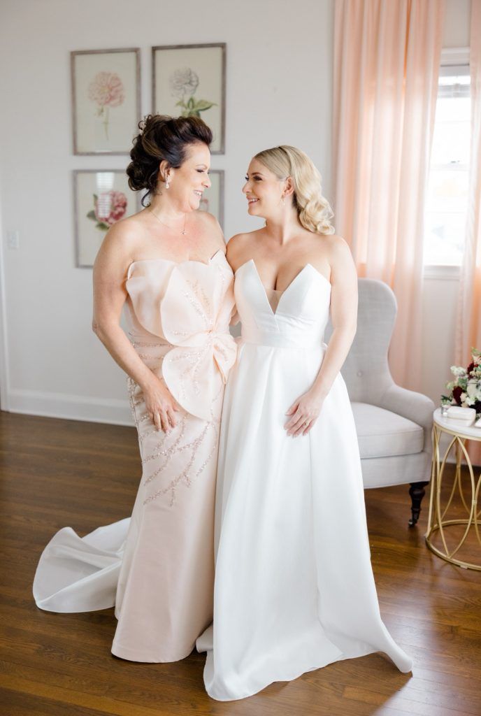 A bride in a white gown smiles at her mother, who wears a blush gown with floral detail, both standing in a softly decorated bridal suite.
