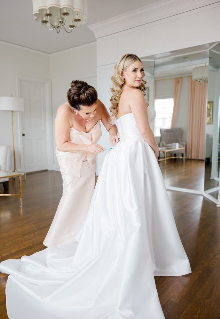 A mother helps button the back of her daughter’s flowing white wedding dress in a bridal suite with mirrors and soft peach curtains.
