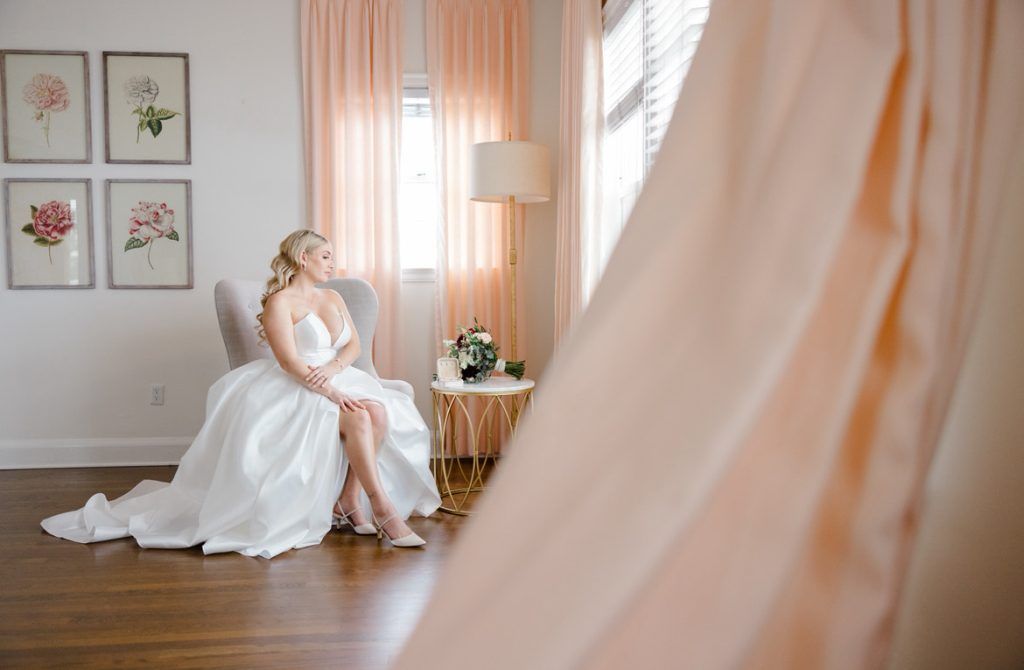 A bride in a white gown sits gracefully on a chair near a window with peach curtains, holding her arms and gazing thoughtfully to the side.