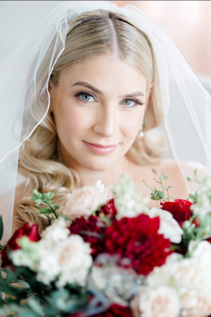 Close-up of a bride in a veil holding red and blush florals, smiling softly with curled hair draped over one shoulder.