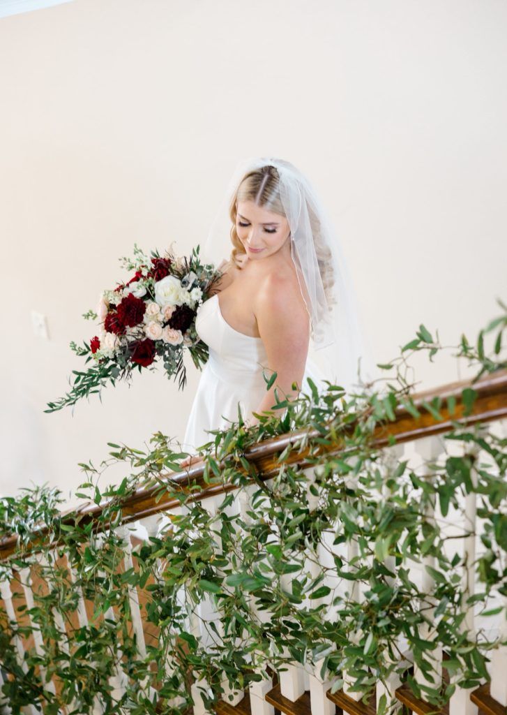 A bride in a strapless gown and veil walks down a greenery-adorned staircase, holding a lush bouquet of red, white, and blush flowers.