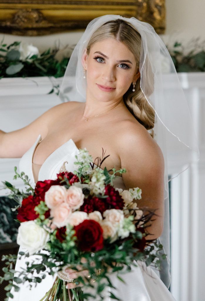 Bride in a white gown and veil poses indoors holding a bouquet of red, blush, and white flowers with greenery accents.