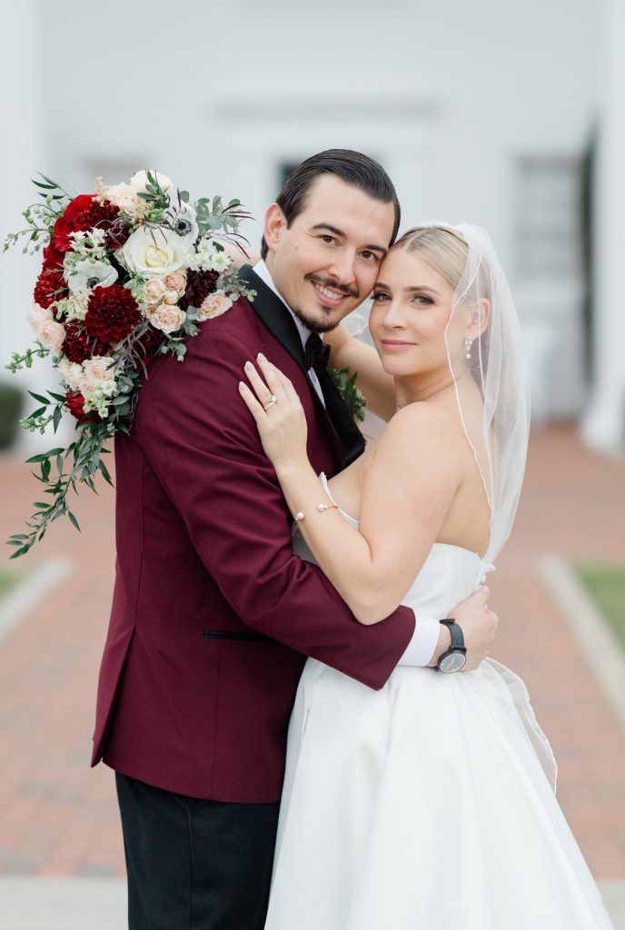Groom in a burgundy tuxedo embraces his bride in a strapless gown with a veil, holding a bouquet of red and blush flowers.