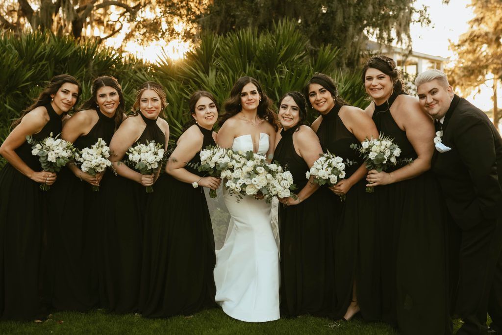 Bride posing with bridal party in black gowns, all holding white rose bouquets at sunset.